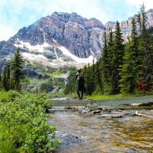 A person with a backpack stands on a wooden footbridge over a rocky stream, facing a large, snow-capped mountain and pine forest.
