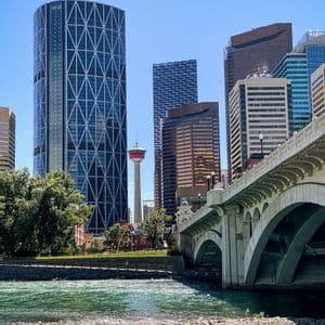 A stone arch bridge crosses a river with a modern city skyline featuring an observation tower in the background under a clear sky.