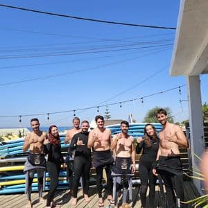 Un gruppo WeRoad in mute fa il segno shaka su un pontile di legno con tavole da surf impilate sullo sfondo.