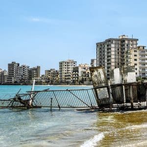 Una recinzione metallica rotta con filo spinato giace parzialmente sommersa nel mare, con edifici cittadini abbandonati che costeggiano la riva sullo sfondo.
