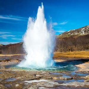 Un geyser projette une haute colonne d'eau et de vapeur depuis un bassin turquoise, avec une colline saupoudrée de neige sous un ciel bleu éclatant.