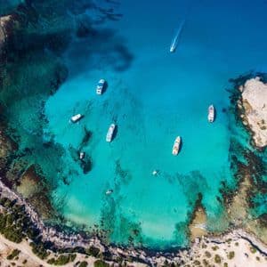 Una vista aerea di diverse barche ancorate in una baia cristallina e turchese vicino a una costa rocciosa e un piccolo isolotto.