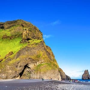 Eine WeRoad Gruppenreise an einem schwarzen Sandstrand am Fuße einer großen, moosbewachsenen Klippe mit aus dem Wasser ragenden Felsnadeln.