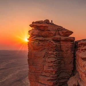 Una persona si trova su un'imponente formazione rocciosa che domina un vasto paesaggio desertico durante un vivace tramonto arancione.