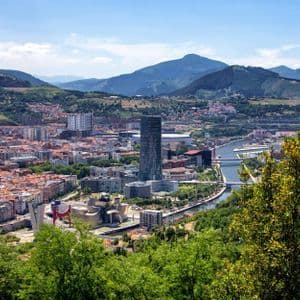 A panoramic view of a city with a winding river, nestled in a valley with green hills and distant mountains.