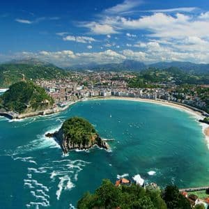 An aerial view of a coastal city along a crescent-shaped bay with a sandy beach, with a small island and green hills.