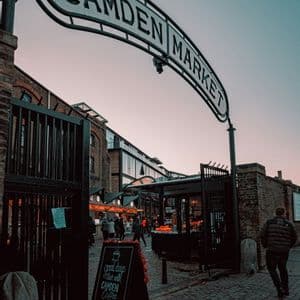 The entrance sign to Camden Market arches over a cobblestone street with people walking below at dusk.