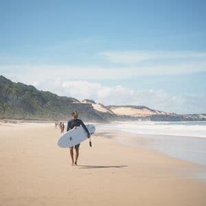 Un surfista in muta cammina su una spiaggia sabbiosa portando una tavola da surf, con onde e palme sullo sfondo.