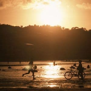 Sagome di persone su una spiaggia bagnata al tramonto, con una persona che corre e un'altra che cammina con una bicicletta in riva al mare.