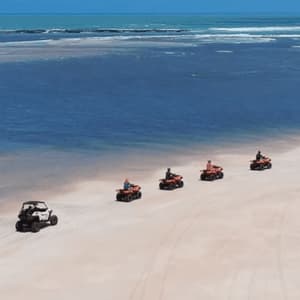 Eine WeRoad-Gruppe fährt mit Quads und einem UTV in einer Reihe entlang eines Sandstrandes neben dem blauen Ozean.