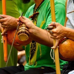 Un primo piano di due persone che suonano berimbau tradizionali e tengono in mano sonagli caxixi.