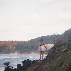 Una donna con una tavola da surf si erge su una scogliera rocciosa che domina una spiaggia con onde marine sotto un cielo nuvoloso.