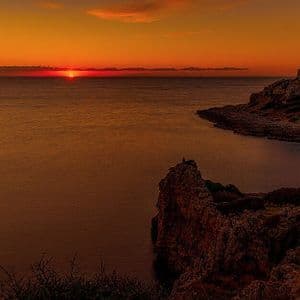 Una scogliera rocciosa con una piccola torre in pietra si staglia contro un tramonto arancione sul mare.