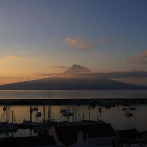 Un porto con barche a vela ormeggiate al tramonto, con un grande vulcano circondato da nuvole stagliato sull'acqua.