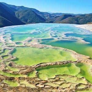 Piscinas minerales en terrazas de verde y turquesa vibrante, con vistas a una vasta cordillera bajo un cielo azul claro.