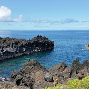 Rocce vulcaniche scure e frastagliate formano una piccola insenatura con acque limpide e turchesi in una giornata di sole lungo la costa.