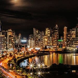 A city skyline of illuminated skyscrapers at night, with light trails on a road next to the water.