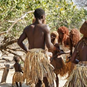 A group of tribal people in traditional attire, including reed skirts, dance while a man plays a drum outdoors.