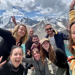 Un voyage de groupe WeRoad sourit pour un selfie avec un arrière-plan panoramique de montagnes enneigées et un lac turquoise.