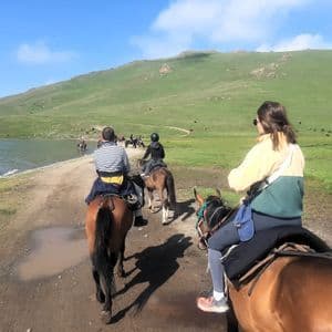 Un voyage de groupe WeRoad à cheval sur un chemin de terre le long d'un lac, avec des collines verdoyantes en arrière-plan.
