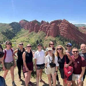 Un voyage de groupe WeRoad prend un selfie devant une formation rocheuse rouge et une vallée verdoyante et arborée sous un ciel bleu clair.