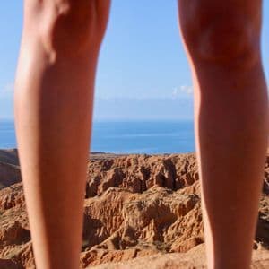 Une vue entre les jambes d'une personne, donnant sur un canyon de roches rouges, un vaste lac bleu et des montagnes lointaines.