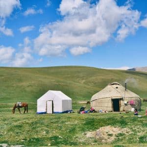Une yourte traditionnelle et une tente blanche sont installées dans une vaste prairie verdoyante, avec des collines ondulantes en arrière-plan, et une famille ainsi qu'un cheval à proximité.