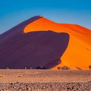 Eine große, orangefarbene Sanddüne wirft einen tiefen Schatten auf sich selbst in einer felsigen Wüstenlandschaft unter einem klaren blauen Himmel.