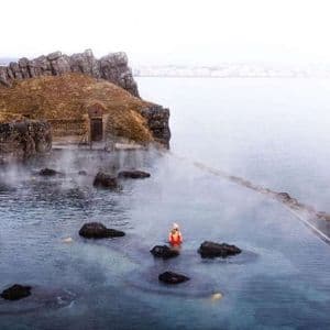 Une personne portant un bonnet et un maillot de bain rouge se détend dans une piscine à débordement fumante intégrée à une côte rocheuse.