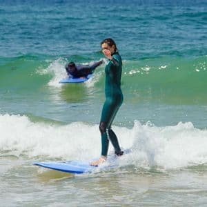 Una donna in muta verde si trova su una tavola da surf blu, cavalcando una piccola onda durante una giornata di sole in un viaggio di gruppo WeRoad.