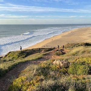 Un gruppo WeRoad si trova su una scogliera erbosa, osservando le onde che si infrangono su un'ampia spiaggia sabbiosa sotto un cielo blu.