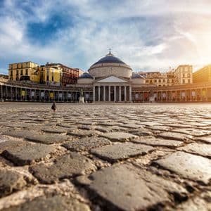 Una veduta dal basso di un piccione che cammina su una vasta piazza acciottolata, di fronte a un grande edificio a cupola con colonne.
