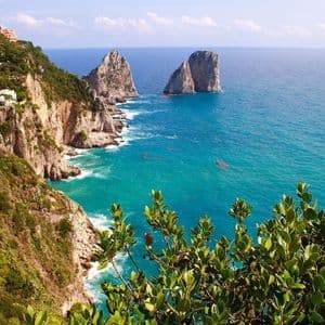 Una vista da una scogliera di due grandi faraglioni in un mare turchese, con vegetazione verde che copre la costa e case costruite sulle colline.