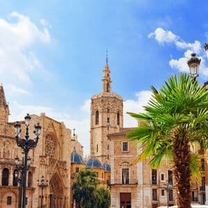 Bâtiments historiques en pierre et un clocher proéminent sur une place de ville avec un palmier sous un ciel ensoleillé et partiellement nuageux.