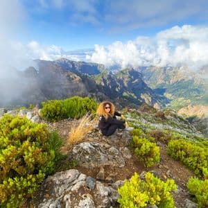 Una mujer con cabello rizado se sienta en la cima rocosa de una montaña, rodeada de arbustos verdes, contemplando un vasto valle montañoso lleno de nubes.