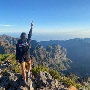 Un líder de grupo de WeRoad, de espaldas a la cámara, de pie en la cima de una montaña rocosa, haciendo el signo de la paz sobre una vasta cadena montañosa.