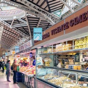 Des gens faisant des achats aux étals de nourriture dans une halle de marché intérieure animée, avec un haut plafond architectural orné.
