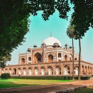Un mausoleo ornato in arenaria rossa con un'ampia cupola bianca è incorniciato da alberi, con un prato verde e un sentiero in primo piano.