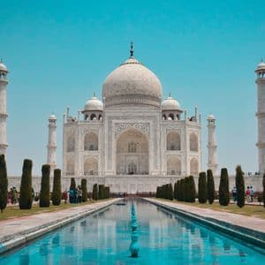 Il Taj Mahal di marmo bianco si riflette in una lunga vasca d'acqua, affiancato da giardini e alberi sotto un cielo azzurro e limpido.