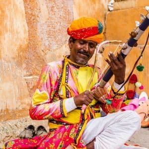 Un uomo con un turbante colorato e abiti tradizionali suona uno strumento a corde seduto accanto a un'esposizione di scarpe.