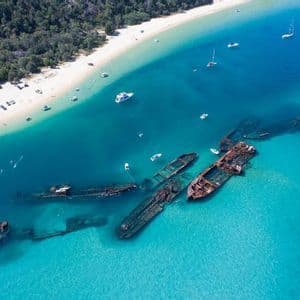 Vue aérienne de plusieurs épaves de navires coulés reposant dans des eaux turquoises peu profondes, près d'une plage de sable bordée d'arbres avec des bateaux.