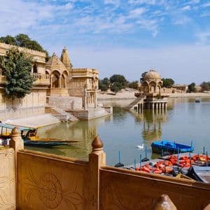 Ornate sandstone temples and pavilions stand on the edge of a calm lake, with colorful boats moored at the ghats under a blue sky.