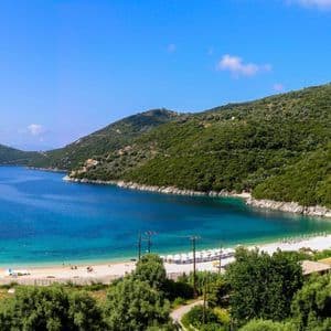 Ein Panoramablick auf eine türkisfarbene Bucht mit einem weißen Sandstrand und einem einsamen Segelboot, umrahmt von bewaldeten Hügeln.