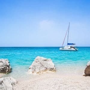 Un catamarano a vela bianco galleggia su acque tranquille e turchesi, visto da una spiaggia di ciottoli con grandi rocce in primo piano.