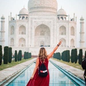 Una mujer con un vestido rojo y una mochila WeRoad posa con el brazo levantado frente al Taj Mahal y su estanque reflectante.