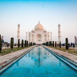 Il Taj Mahal di marmo bianco si riflette in una lunga e tranquilla piscina turchese, affiancata da giardini con cipressi sotto un cielo sereno.