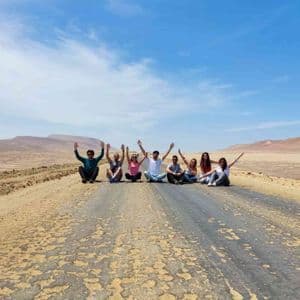 Un grupo de viaje de WeRoad se alinea en una carretera desértica con los brazos en alto bajo un cielo azul brillante.