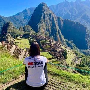 Una mujer con una camiseta de "WeRoad Group Leader" se sienta en una colina cubierta de hierba con vistas a ruinas antiguas anidadas entre escarpadas montañas verdes.