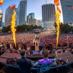 Due DJ si esibiscono su un palco per una folla immensa a un festival all'aperto con fuochi d'artificio che sparano fiamme, sullo sfondo dello skyline di una città.
