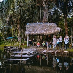 Un viaje en grupo de WeRoad posa en la orilla de un río en la jungla junto a un muelle de madera y una cabaña con techo de paja, rodeados de palmeras.